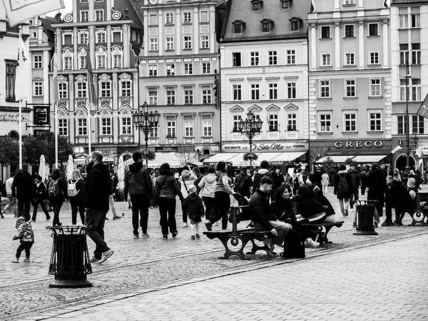 A bustling European city square filled with people, showcasing historical architecture in grayscale.