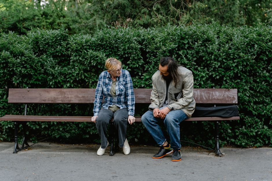 Two adults having a conversation on a wooden park bench surrounded by greenery.