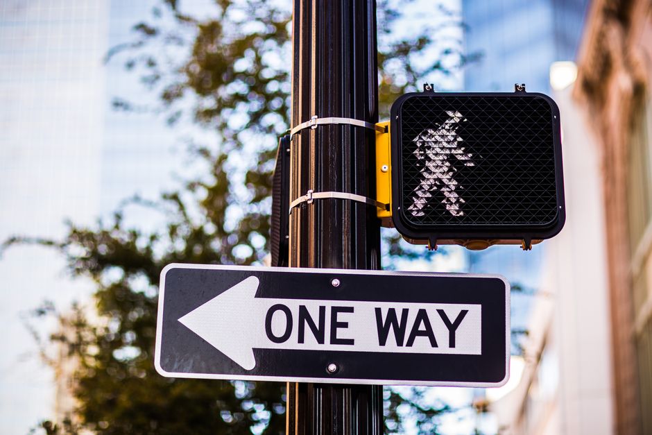 Urban one way street sign and pedestrian crossing light, daytime city scene.